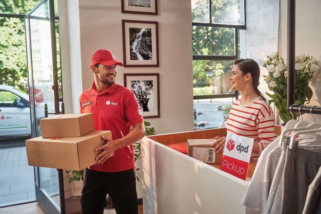 Uśmiechnięty kurier DPD w czerwonej czapce i koszulce przekazuje paczki klientce w punkcie odbioru DPD Pickup w jasnym wnętrzu sklepu.