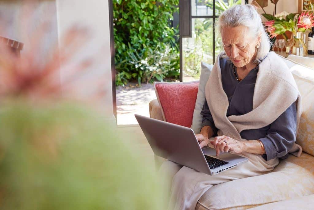 Starsza kobieta w swetrze używa laptopa siedząc na kanapie w jasnym, przytulnym salonie.