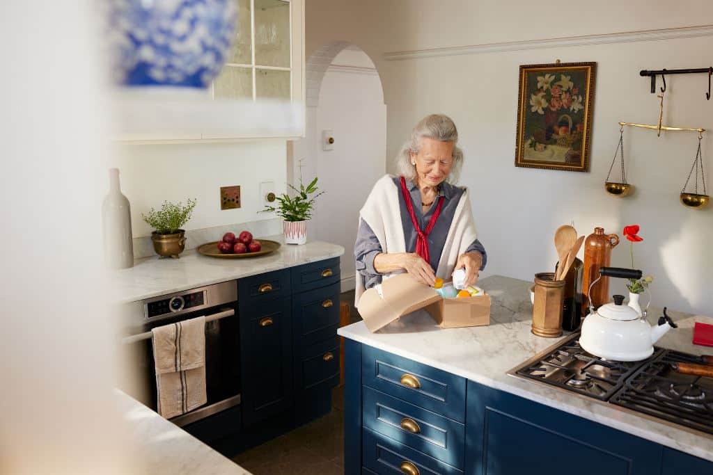 Starsza kobieta otwiera kartonowe pudełko w jasnej kuchni o klasycznym wystroju.