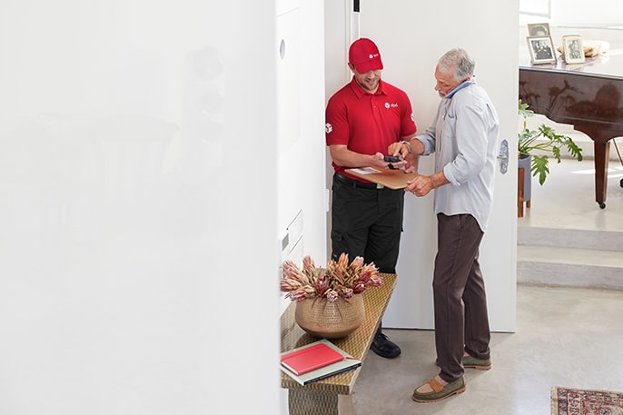 Kurier DPD w czerwonej koszulce i czapce dostarcza przesyłkę starszemu mężczyźnie w domowym zaciszu.